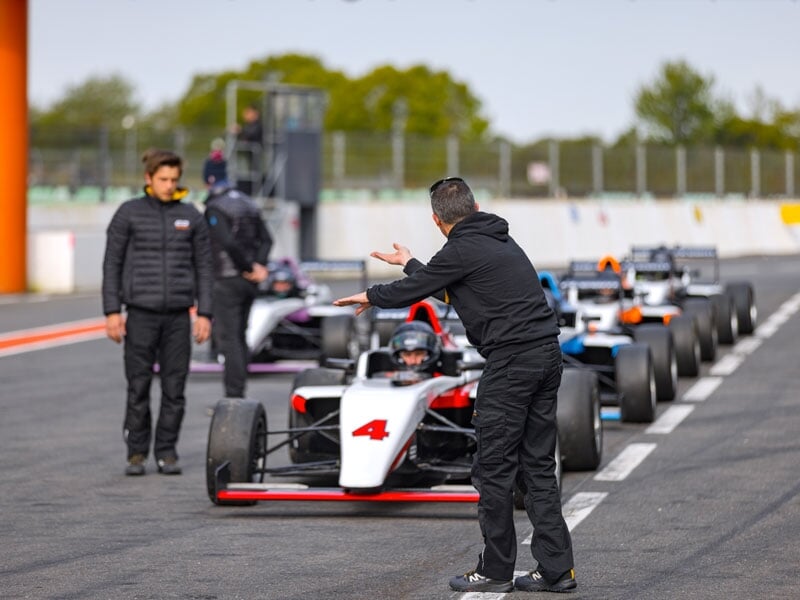 Instructeur donnant des indications à un pilote en monoplace de course blanche et rouge sur la piste, avec plusieurs voitures alignées derrière