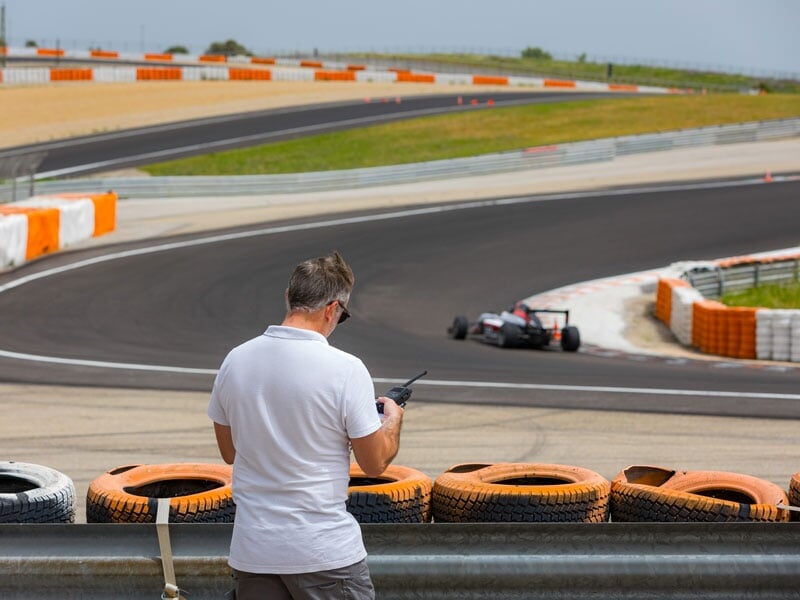 Homme en polo blanc observant une monoplace en pleine course depuis le bord de la piste, équipé d'une radio, près d'une barrière de pneus orange et blanche