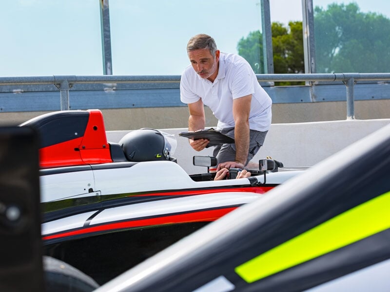 Instructeur en polo blanc utilisant une tablette pour encadrer un pilote dans une monoplace rouge et blanche, positionnée dans la voie des stands