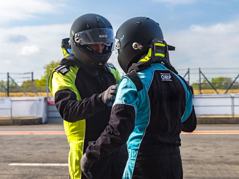 Deux pilotes en combinaisons et casques noirs discutant près de la piste, l'un portant des détails jaunes fluo, l'autre bleu turquoise, dans un contexte d'école de pilotage