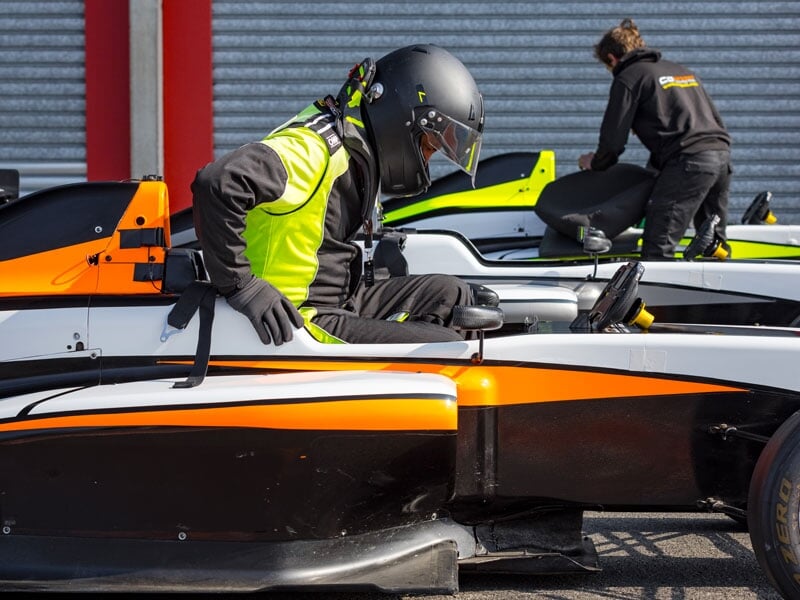 Pilote en combinaison noire et jaune fluo s'installant dans un cockpit de monoplace blanche et orange, prêt pour une session sur circuit automobile