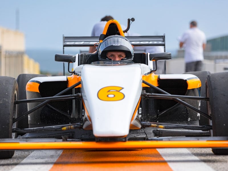 Pilote au volant d'une monoplace blanche et orange, numéro 6 visible sur le capot, positionné sur la ligne de départ dans la voie des stands, entouré par des membres de l'équipe en arrière-plan