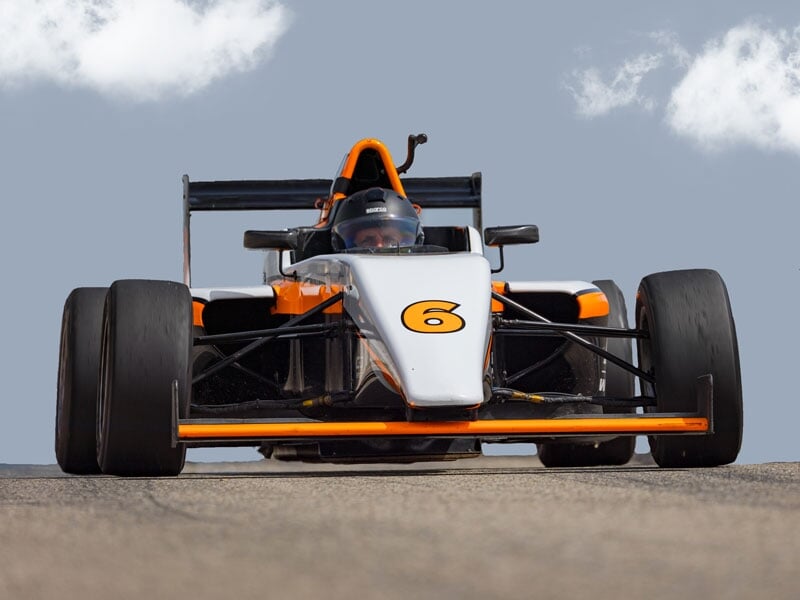 Monoplace blanche et orange numéro 6 en pleine action sur circuit, vue de face avec le pilote visible sous son casque, ciel partiellement nuageux en arrière-plan