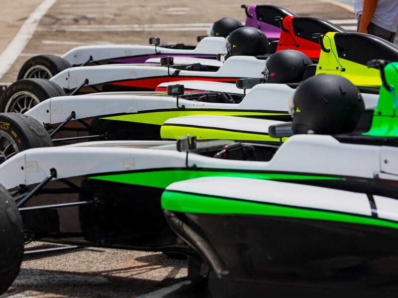 Alignement de monoplaces aux couleurs vives (vert, jaune, rouge, violet) dans la voie des stands, prêtes pour une session, avec casques noirs posés sur les cockpits
