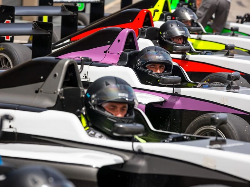 Pilotes casqués dans des monoplaces colorées (violet, rouge, vert), alignés sur la ligne de départ pour une compétition.