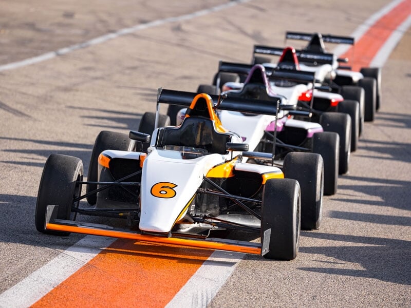 Monoplace de course blanche et orange avec le numéro 6 en tête d’un peloton sur une piste asphaltée, soulignée par une ligne orange, illustrant une compétition automobile.