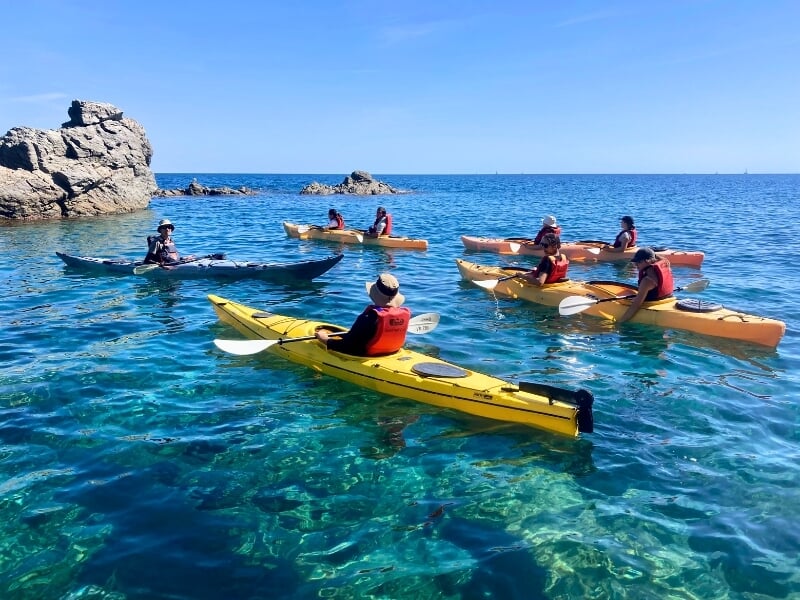 Groupe de personnes faisant du kayak en mer par une journée ensoleillée, avec des rochers émergeant de l'eau claire et un ciel bleu en arrière-plan.