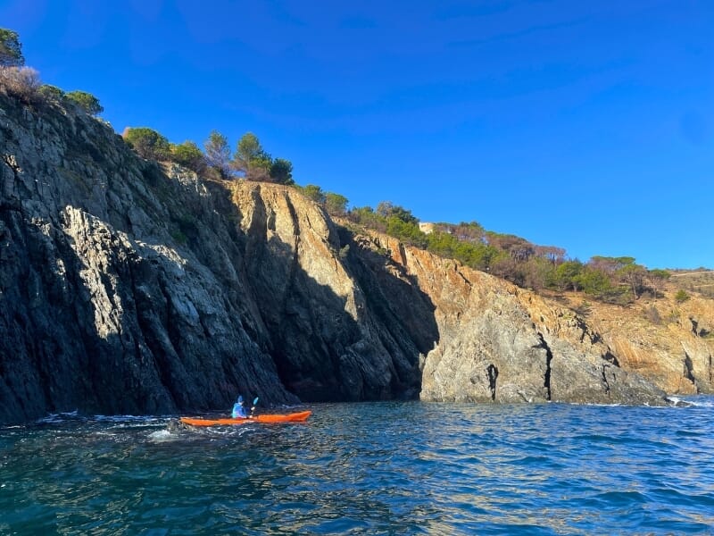 Personne en kayak orange naviguant seul le long d'une côte escarpée avec des falaises abruptes et de la végétation en surplomb, sous un ciel clair