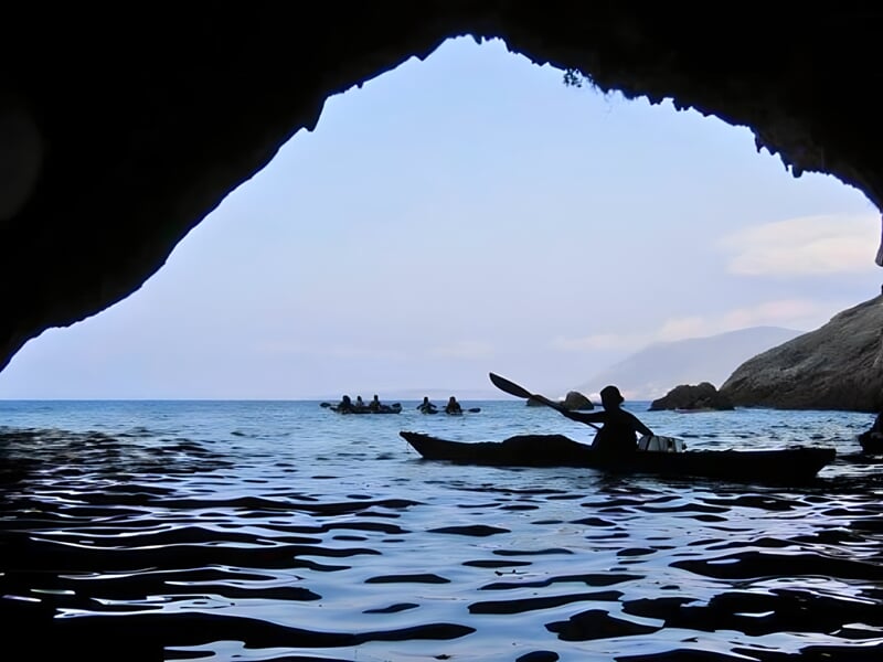 Vue depuis l'intérieur d'une grotte marine sombre avec l'ouverture révélant un kayakiste et d'autres kayaks en arrière-plan sur la mer calme