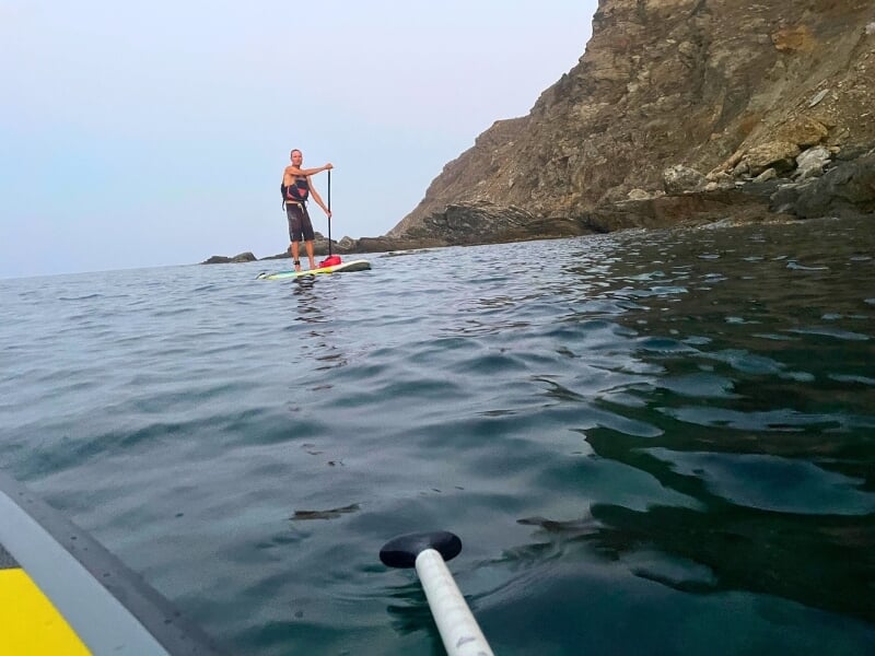 Un homme sur une planche de paddle en mer, pagayant près d'une côte rocheuse, avec la perspective de la caméra depuis une autre planche