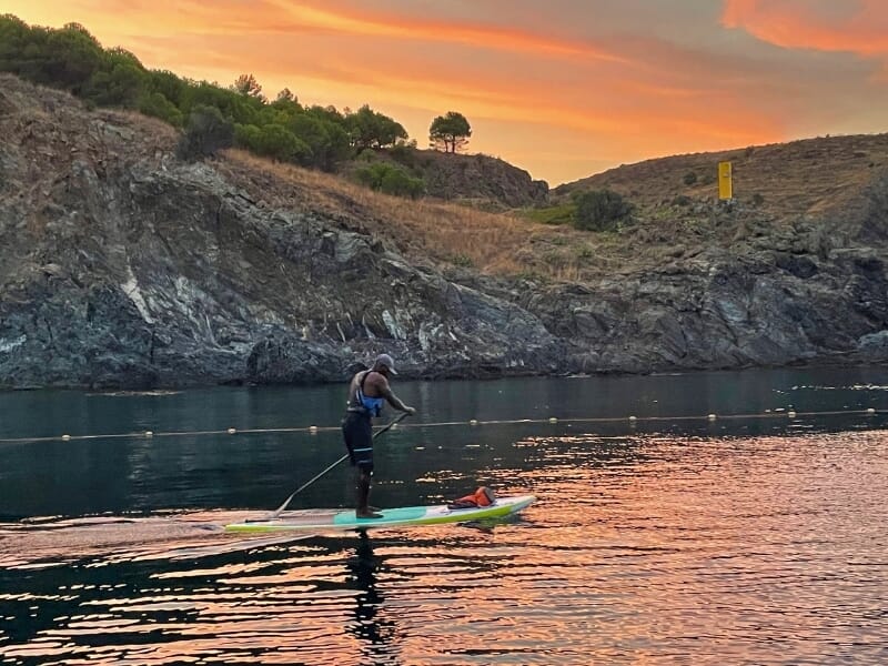 Paddle au coucher du soleil avec un homme sur une planche en mer, contre un ciel orange vibrant et des falaises sombres en arrière-plan.