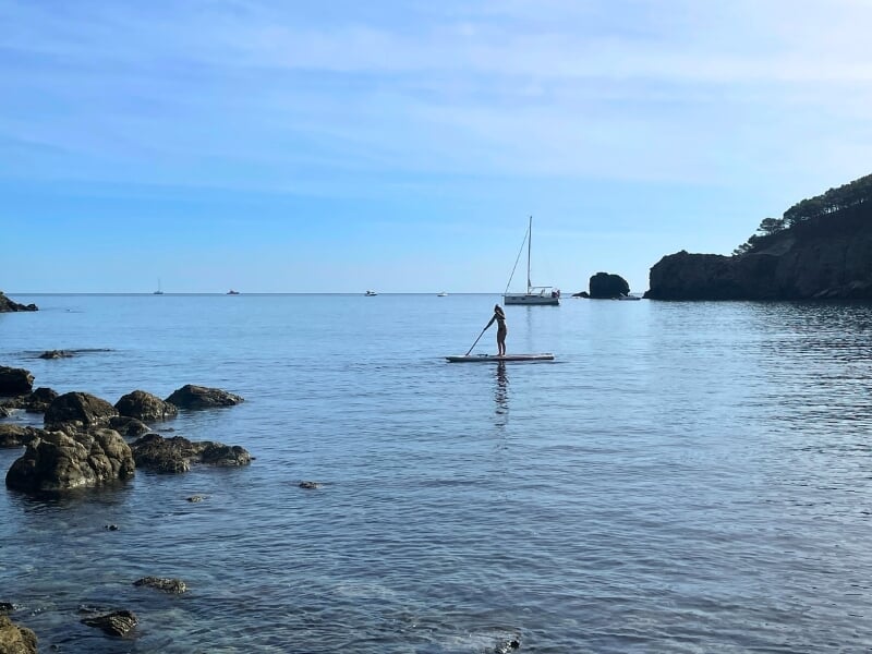 Vue tranquille d'une personne faisant du paddleboard sur une eau calme, avec des bateaux au loin et une côte rocheuse sous un ciel bleu clair.