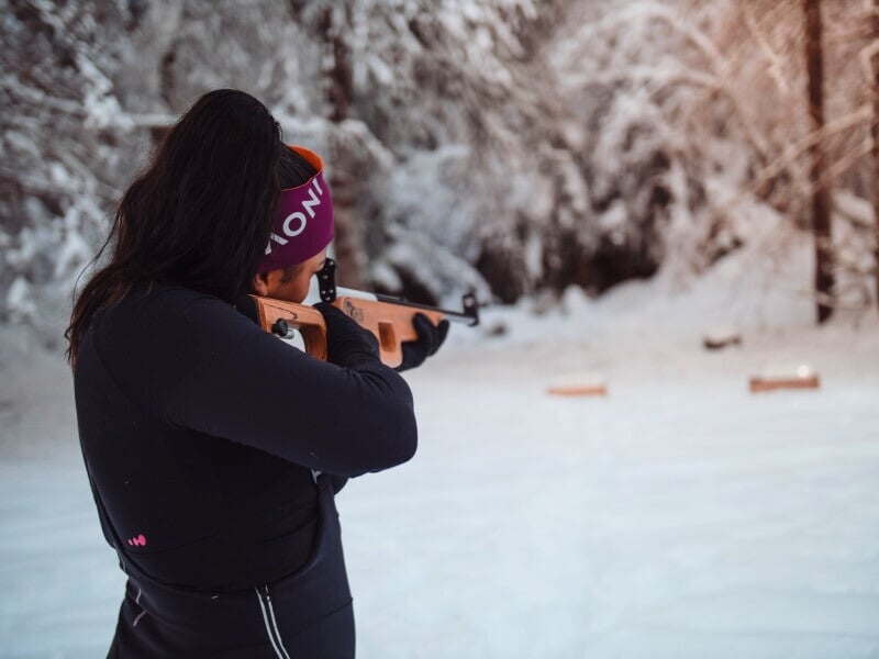 Une femme en tenue de sport d'hiver, portant un bandeau violet, vise avec un fusil de biathlon dans un paysage enneigé.