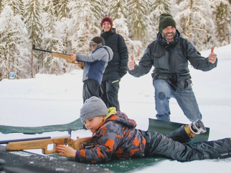 Une famille de quatre personnes s'amusant dans la neige ; un enfant est allongé visant avec un fusil de biathlon, un autre tient un fusil en arrière-plan, et deux adultes sourient et font des pouces l