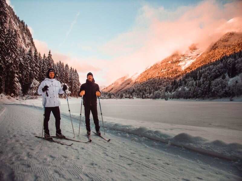 Deux hommes en tenue de ski de fond, l'un en blanc et l'autre en noir, skiant sur une piste enneigée avec une forêt et une montagne illuminée par un coucher de soleil en arrière-plan.