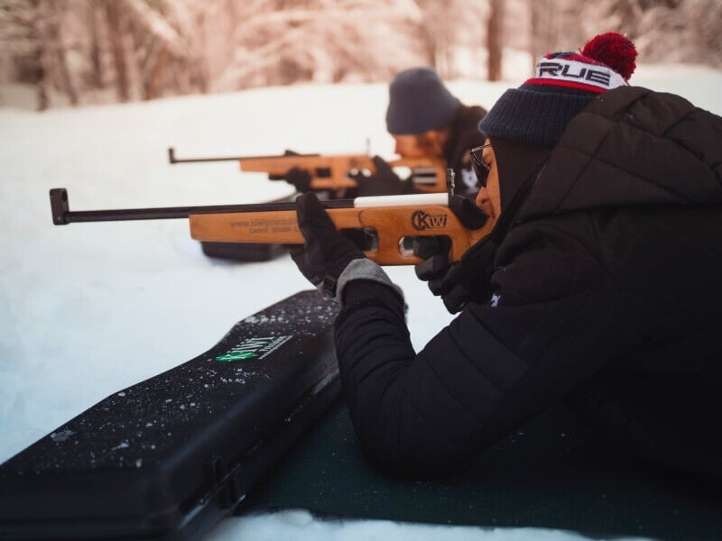 Un homme allongé sur le ventre dans la neige, visant avec un fusil de biathlon, portant un bonnet et des gants, avec une mallette noire à côté de lui.
