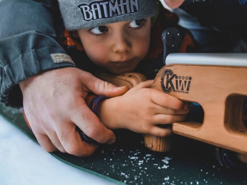 Gros plan sur les mains d'un enfant tenant un fusil de biathlon, avec une aide adulte, apprenant à viser sur une surface enneigée.