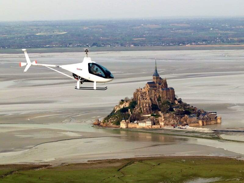 Un hélicoptère ULM CH-77 vole au-dessus de la Baie du Mont-Saint-Michel avec l'abbaye en second plan
