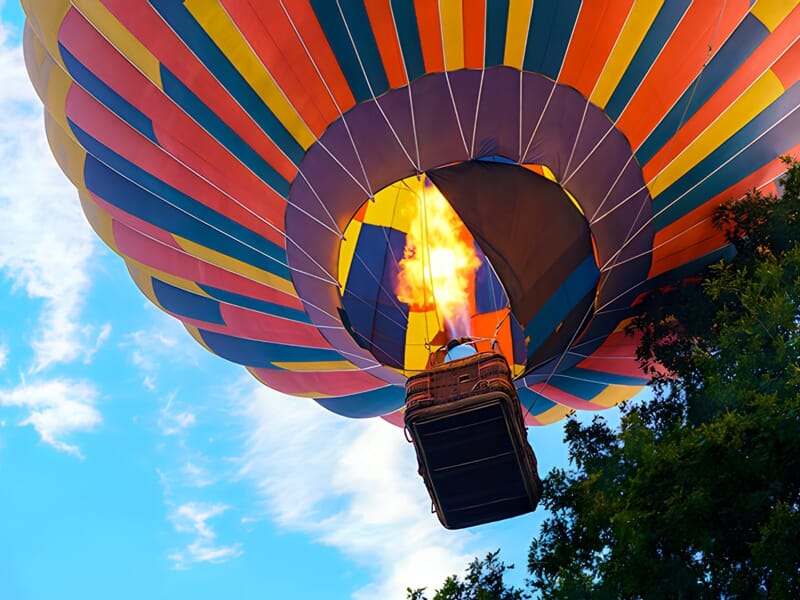 Montgolfière multicolore en vol vue du dessous, avec des flammes visibles dans le brûleur, contre un ciel bleu avec quelques nuages, capturant la dynamique et l'excitation d'un vol en montgolfière