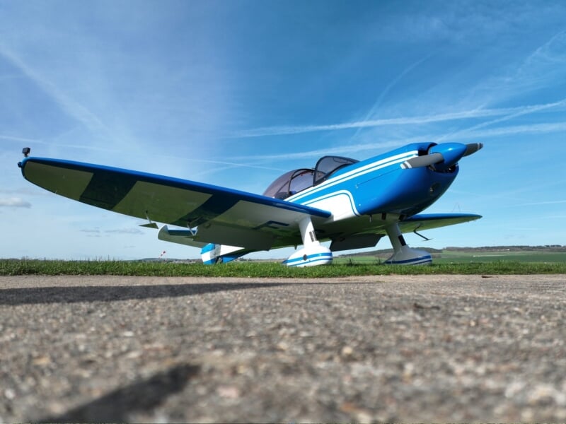 Avion monomoteur bleu et blanc sur une surface en béton, avec un horizon dégagé et un ciel bleu clair.