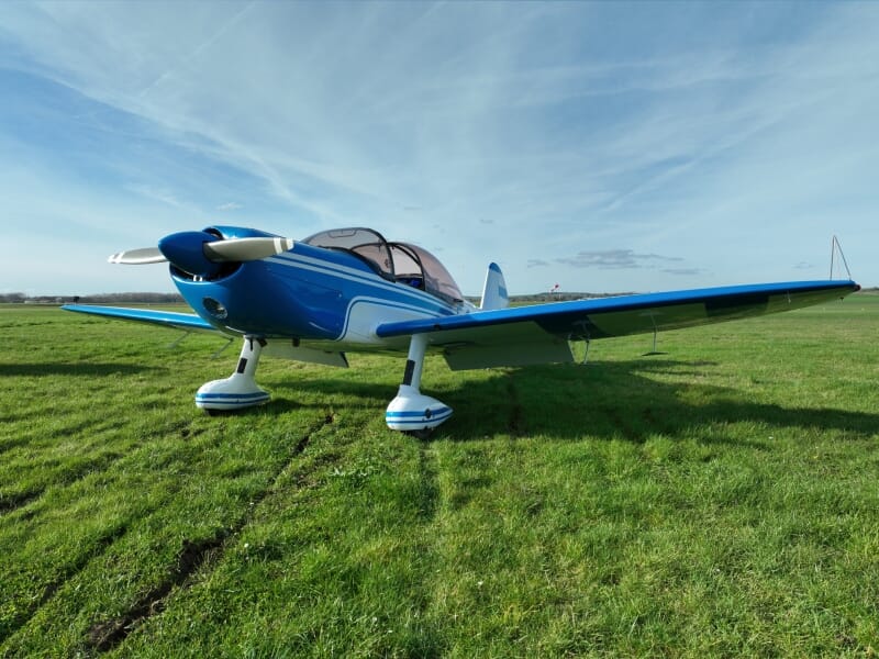 Avion monomoteur bleu et blanc garé sur une pelouse verte sous un ciel bleu clair avec quelques nuages. La cabine transparente et le train d'atterrissage robuste sont bien visibles.