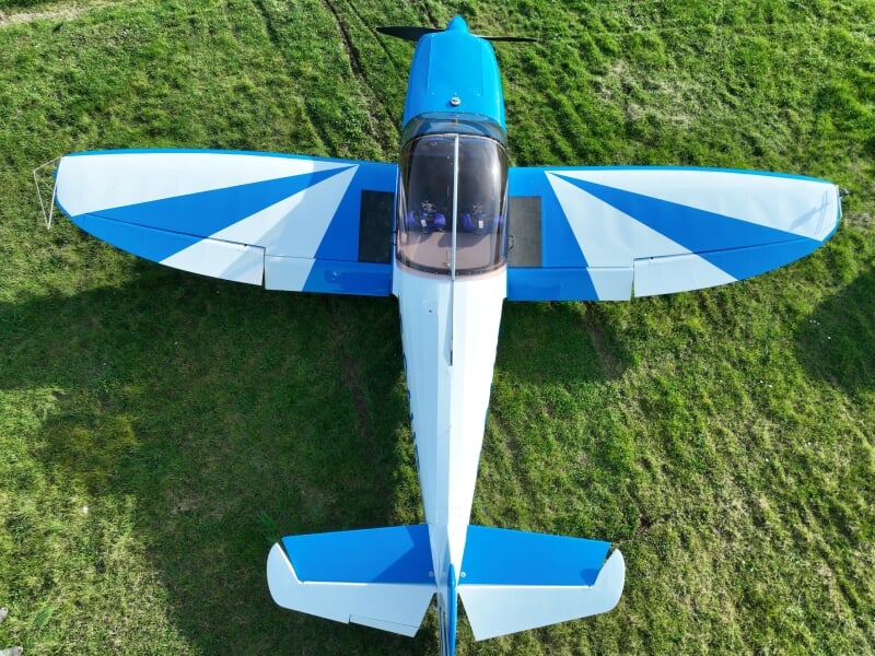 Vue aérienne d'un avion de voltige bleu et blanc stationné sur une pelouse verte. Les ailes arborent un motif géométrique en étoile, accentuant son design dynamique. Le cockpit vitré laisse entrevoir