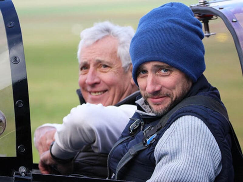 Deux hommes souriants assis dans le cockpit d'un petit avion, l'un portant un bonnet bleu et l'autre avec des cheveux gris.