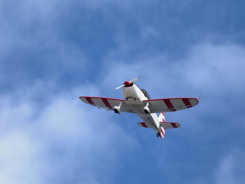 Petit avion de voltige blanc avec des rayures rouges en vol contre un ciel bleu avec quelques nuages.