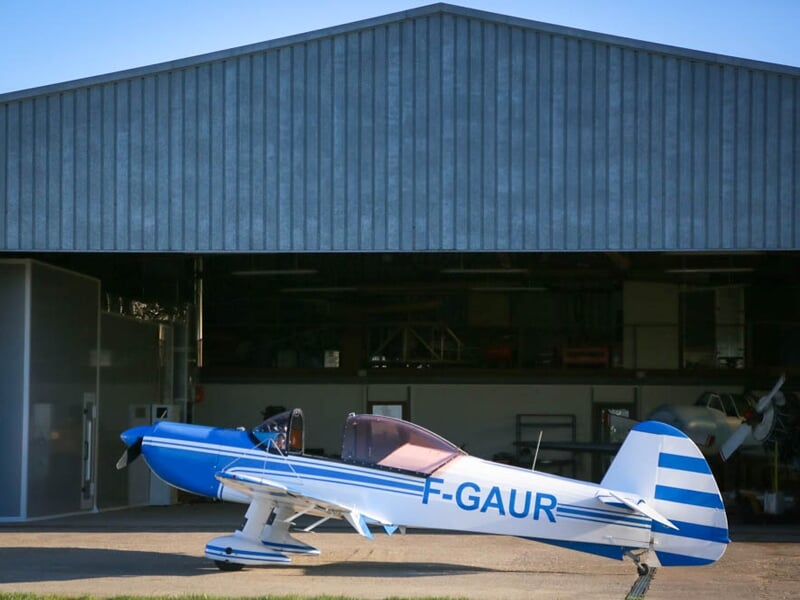 Avion de voltige bleu et blanc, immatriculé F-GAUR, stationné devant un hangar métallique ouvert. Son design aérodynamique et son cockpit ouvert laissent présager des performances acrobatiques.