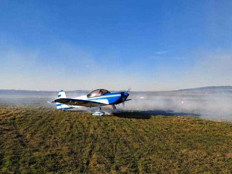 Avion de voltige sur une piste en herbe, moteur en marche, dégageant de la fumée. L’arrière-plan montre un paysage vallonné sous un ciel clair, idéal pour une démonstration aérienne.