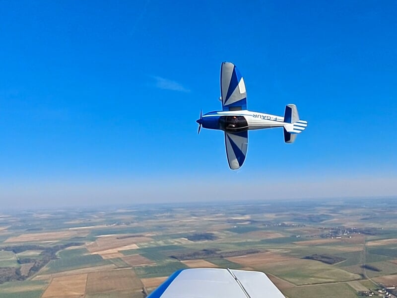 Avion de voltige effectuant une figure acrobatique en vol, entièrement inversé au-dessus d’un paysage rural. L'immatriculation F-GAUR est bien visible, soulignant la précision et l’adrénaline du pilot