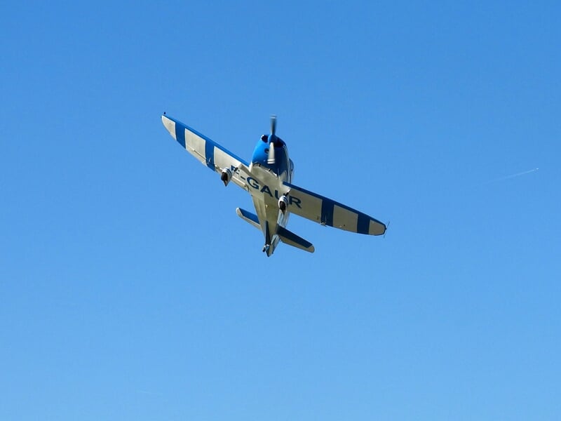 Avion de voltige bleu et blanc en plein vol sous un ciel dégagé, capturé en contre-plongée. L'hélice en mouvement et l'immatriculation visible sur le fuselage ajoutent du dynamisme à l'image.