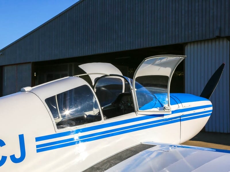 Gros plan sur un avion léger stationné devant un hangar, avec ses portes papillon ouvertes. La lumière du soleil se reflète sur le fuselage blanc aux lignes bleues, mettant en valeur son design.