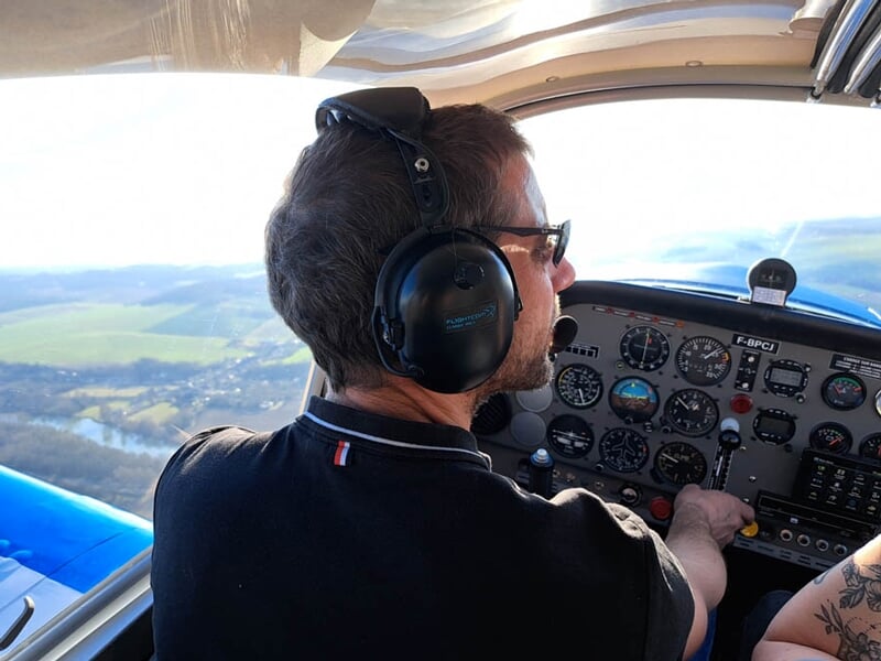 Pilote aux commandes d’un avion léger, portant un casque audio et concentré sur les instruments de vol. La vue extérieure montre un paysage vallonné sous un ciel dégagé.