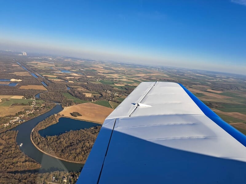 Vue aérienne depuis l’avion, capturant l’aile blanche et bleue avec en arrière-plan un paysage de rivières sinueuses, forêts et terres agricoles. L'horizon s’étend sous une lumière parfaite pour un vo