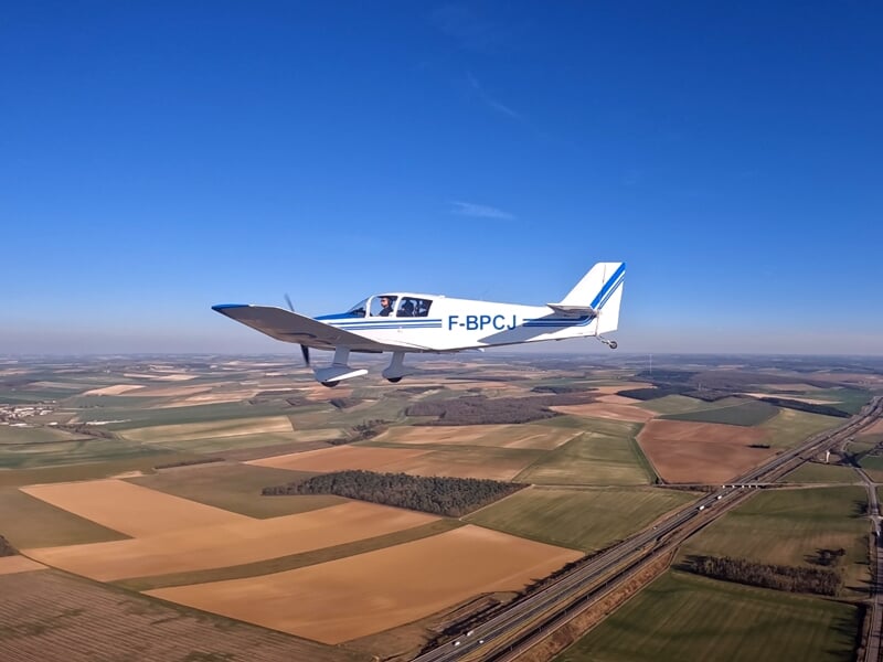 Avion léger en vol au-dessus d’un paysage rural avec champs et routes. Le ciel dégagé et la vue panoramique offrent des conditions idéales pour un vol paisible.
