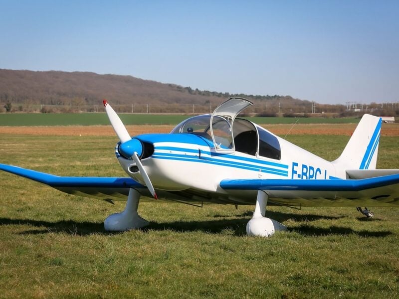 Petit avion blanc et bleu stationné sur une pelouse verte avec une colline boisée en arrière-plan.