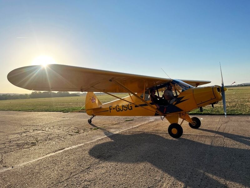 Avion jaune au sol sur une piste en béton avec le soleil brillant à l'arrière et un pilote à bord.