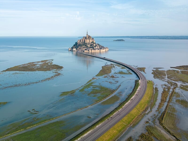 Vue aérienne d'un vol en hélicoptère au-dessus du Mont-Saint-Michel avec sa célèbre abbaye, entouré par la mer et relié par une route sinueuse.