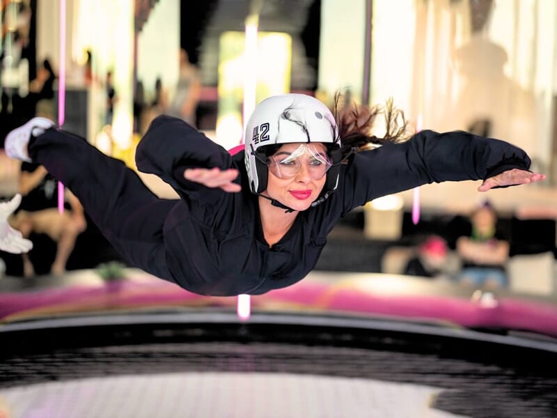 Femme en combinaison de vol noire faisant une expérience de chute libre indoor avec un casque et des lunettes de protection