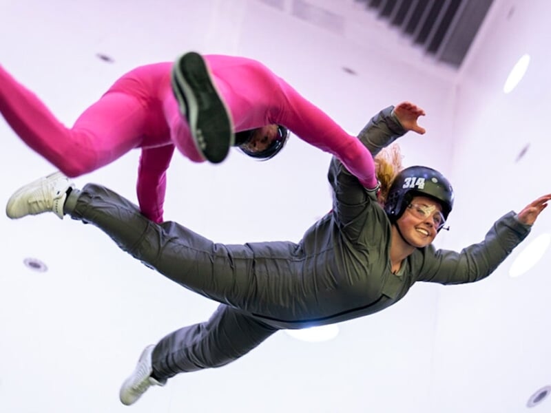 Jeune femme en combinaison de vol rose en position de vol avec l'assistance d'un instructeur en soufflerie intérieure