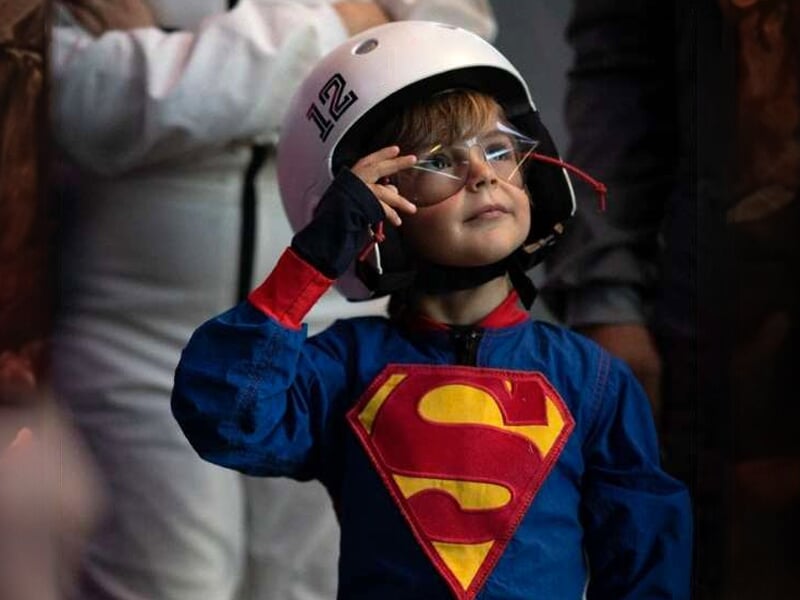 Enfant en costume de Superman avec un casque de vol, faisant un geste de salut dans une soufflerie intérieure
