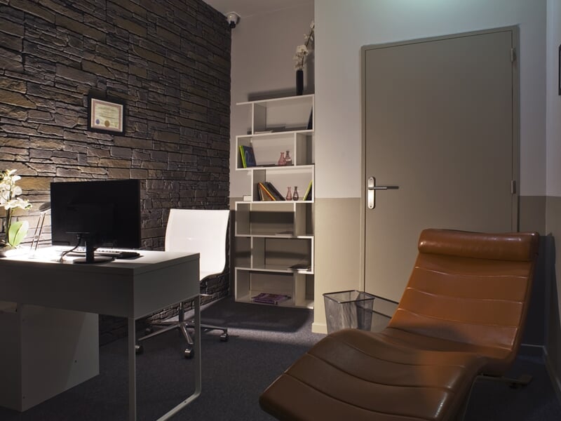 Bureau moderne avec chaise en cuir marron, ordinateur sur table blanche et étagères avec livres dans une pièce claire.