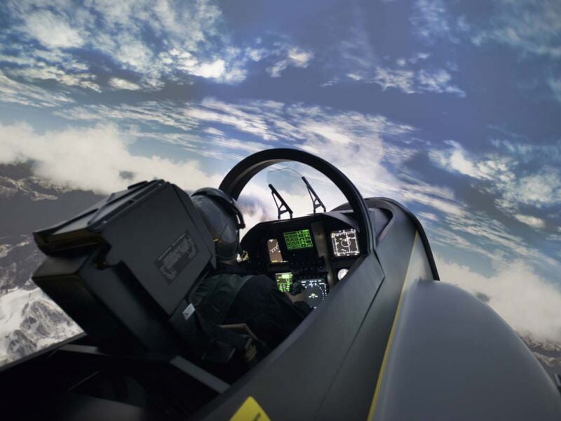 Pilote dans un simulateur d'avion avec casque noir, volant et instruments de bord visibles sous un ciel bleu nuageux.