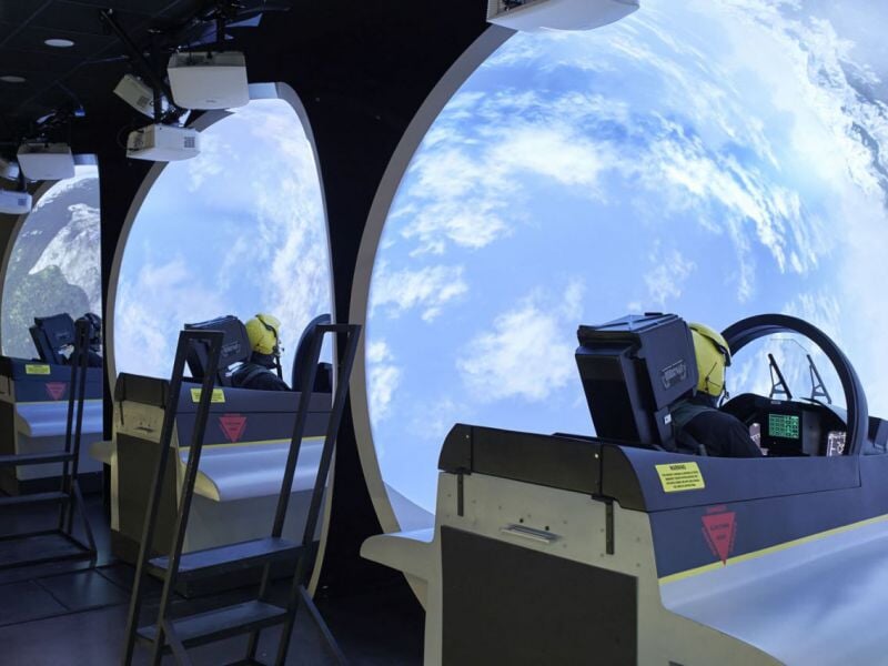 Salle avec plusieurs simulateurs de pilotage d'avion alignés face à un écran circulaire affichant un paysage aérien.
