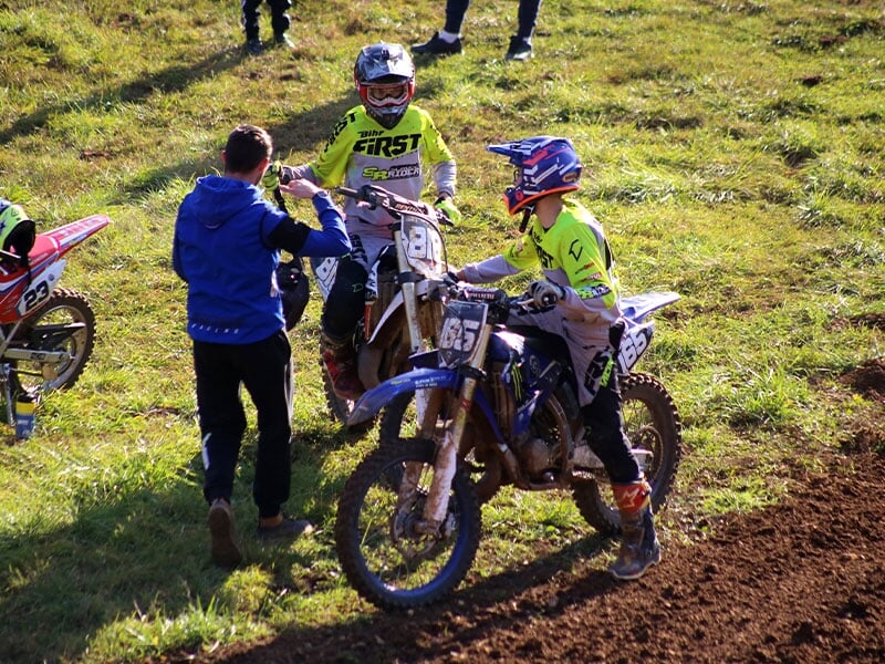 Échange entre un entraîneur en veste bleue et pilotes de motocross, préparation avant la course, motos en file, terrain herbeux