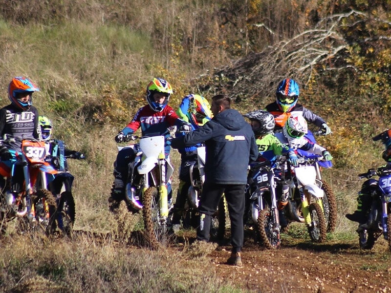 Discussion technique entre entraîneur en noir et pilotes de motocross alignés sur un terrain vallonné, sous un ciel clair