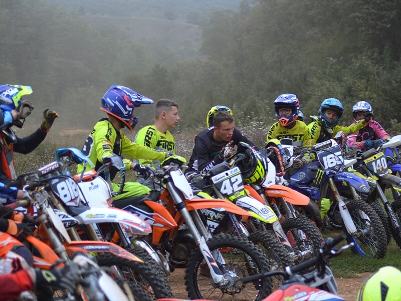 Groupe de pilotes de motocross en tenue de course écoutant les consignes, moto variées en couleur, jour brumeux, nature en fond