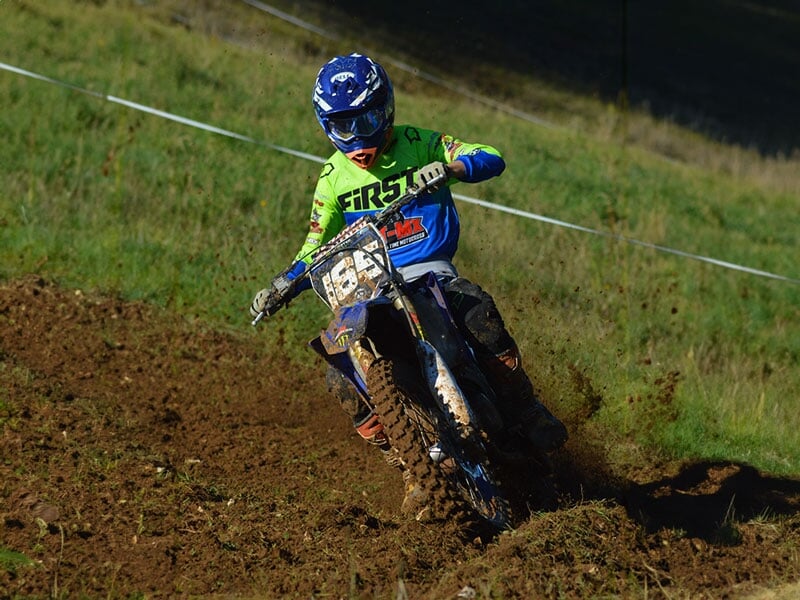 Pilote de motocross vêtu de bleu et de vert fluo manœuvrant avec adresse sa moto, éclaboussures de boue, concentration intense, clôture blanche et verdure en arrière-plan