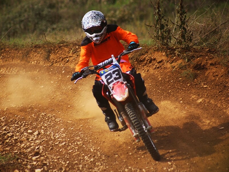 Motard en tenue orange roulant à pleine vitesse sur une piste de terre, poussière en suspension, action dynamique, nature en toile de fond